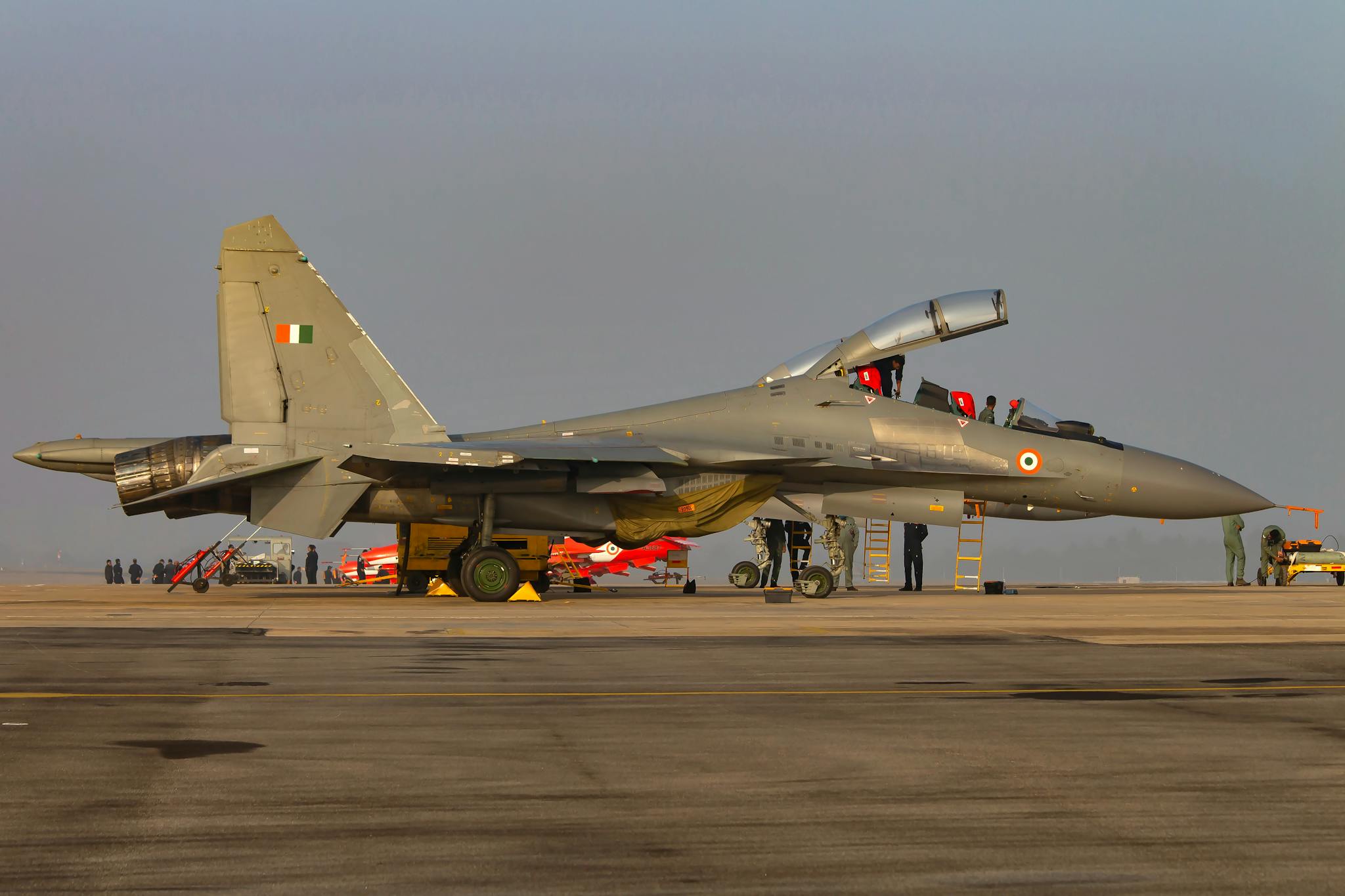 A military jet with open cockpit on a tarmac, surrounded by personnel and equipment.