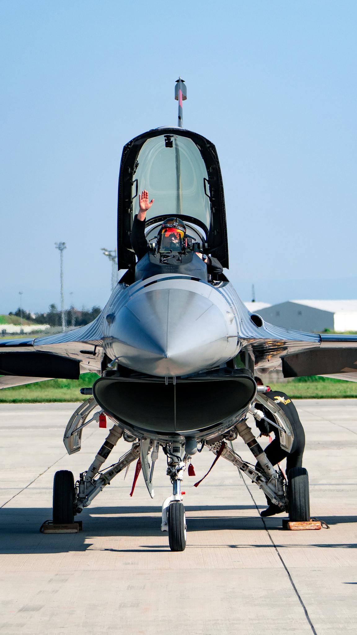 Air force pilot waving from a parked jet on a runway, under clear skies.